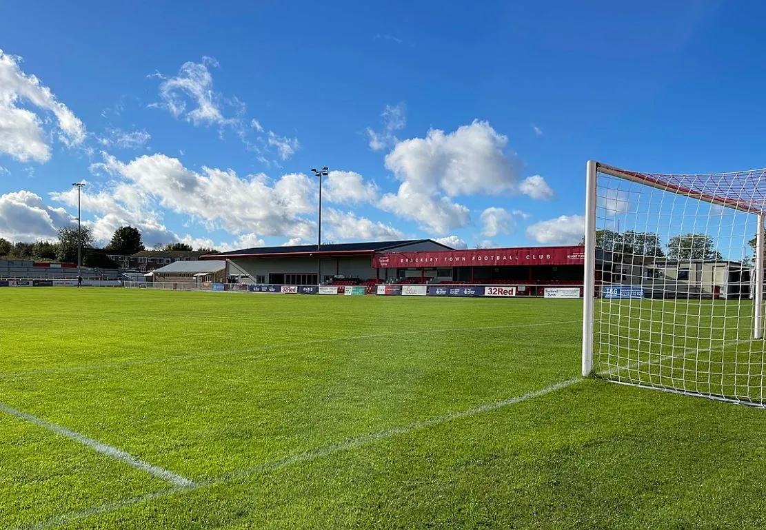 Khán đài chính sân St James Park Brackley, với hàng ghế và mái che đặc trưng của sân bóng đá nhỏ ở Anh.
