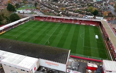 Toàn cảnh sân vận động Whaddon Road của Cheltenham Town FC nhìn từ trên cao, cho thấy cấu trúc độc đáo của một sân bóng hạng dưới Anh.